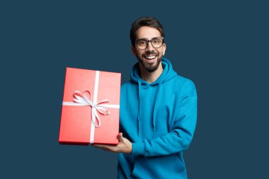 Gözlüklü ve mavi kapüşonlu genç bir adam büyük kırmızı bir hediye kutusu sunar. Beyaz bir kurdeleyle bağlanmış, mavi arka planda heyecan ve neşe sergiliyor..