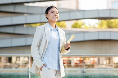 Hafif takım elbiseli profesyonel bir Asyalı kadın elinde bir klasör tutarken gülümsüyor. Kendine güveni etrafındaki şehrin dinamik atmosferine katkı sağlıyor..