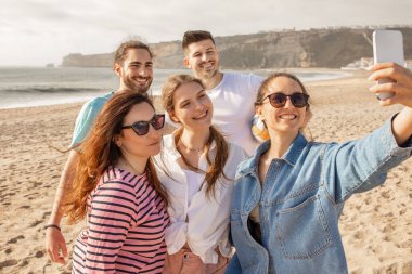 Bir grup arkadaş kumlu bir sahilde selfie çekerken mutlu bir şekilde gülümsüyor. Güneş arka planda batıyor, neşeli toplantının üzerine altın bir ışık saçıyor..
