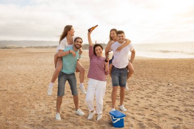 Bir aile eğlenceli bir plaj gününün tadını çıkarırken, diğer üyeler bunu içerek kutlarken iki üye de sırtına biniyor. Güneş kıyı şeridinde batıyor, neşeli bir atmosfer yaratıyor..