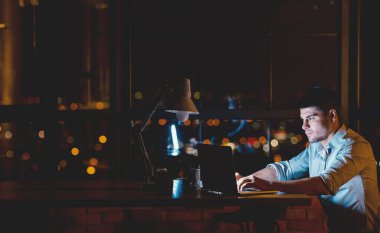 Entrepreneur Guy Working On Laptop Creating Business Startup Overnight Sitting In Modern Office Near Panoramic Window. Free Space For Text