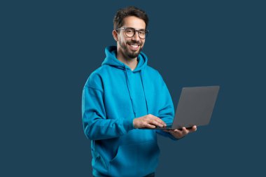 Genç bir adam mavi kapüşonlu bir laptop kullanırken gülümsüyor. Sağlam bir arka plana karşı duruyor, çalışırken rahat ve meşgul bir tutum sergiliyor..