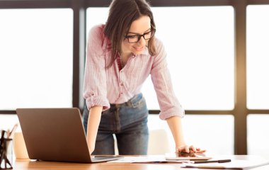 Happy female student standing at desk using digital tablet, touching screen and typing on laptop, copyspace