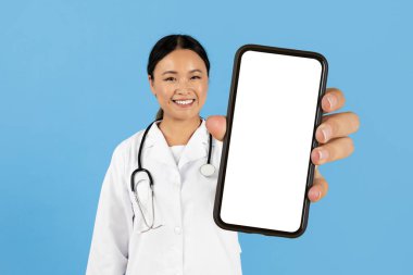 Sağlık çalışanı Asyalı bir kadın mavi bir arka plana karşı duruyor, elinde boş bir ekranla akıllı telefonu tutarken içten bir gülümseme, teletıp veya dijital sağlık seçeneklerini sembolize ediyor..
