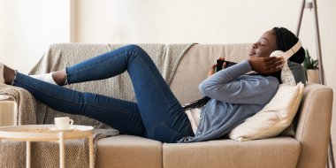 Siyah kız kablosuz kulaklıklarla müzikten hoşlanıyor, kanepeye uzanmış, gözleri kapalı.