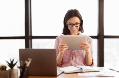 İnternette sörf yapmak. Gözlüklü genç sekreter dijital tablet kullanıyor ve gülümsüyor, modern ofiste masada oturuyor.