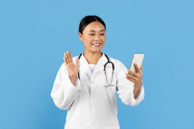 Beyaz önlük ve steteskop giyen gülümseyen Asyalı bir kadın doktor, parlak mavi bir ortamda hastayla uzaktan yakından ilgilenirken akıllı telefonuna el sallayarak sanal bir konsültasyon gerçekleştiriyor..