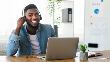 Dizüstü bilgisayarda çalışan, kulaklıkla müzik dinleyen, iş yerinde oturan gülümseyen Afro-Amerikan çalışanı.