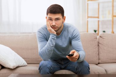 Bored Millennial Guy Switching Channels Watching TV Sitting On Couch At Home. Boring Weekend. Selective Focus