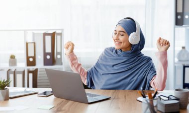 İşteki ruh halini arttırır. Başörtülü neşeli Arap kadın ofiste kablosuz kulaklıkla müzik dinliyor ve masada otururken dans ediyor, panorama
