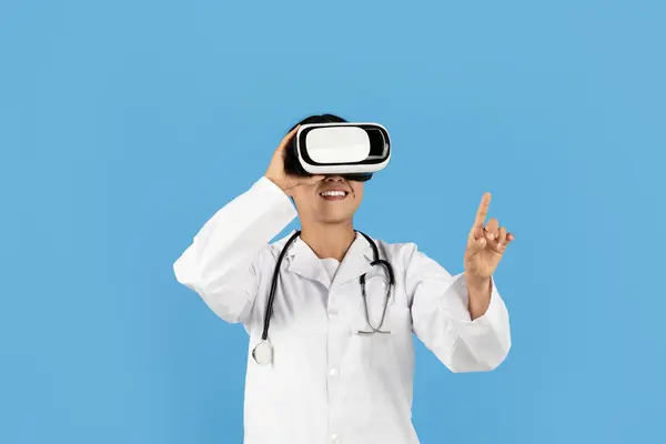 stock image A healthcare professional Asian lady wearing a lab coat and stethoscope engages with a virtual reality headset, smiling while pointing, indicating an immersive experience in a clinical environment.