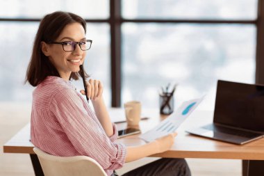 Cheerful Young Woman Sitting At Her Desk With Papers, Posing And Smiling At Camera clipart