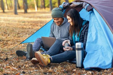 Genç adam ve kadın ormanda kamp yapıyor, dizüstü bilgisayar kullanıyor ve çay içiyor, boşluğu kopyalıyor.