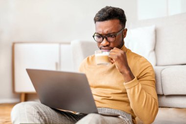 Ciddi bir Afrikalı Amerikalı adam laptopta haber okuyor ve evde yerde oturmuş kahve içiyor. Seçici Odaklanma