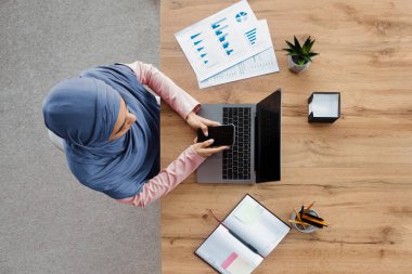 İş İletişimi. Modern ofiste masa başında otururken akıllı telefon kullanan ve dizüstü bilgisayarda çalışan Arapça kadın çalışanın en iyi görünümü