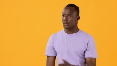 A young man is standing in a relaxed posture, using hand gestures to express his thoughts. He is wearing a light purple t-shirt, with a vibrant orange backdrop enhancing the moment.