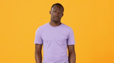 Mor t-shirt giyen genç bir adam, parlak sarı bir zemine karşı duruyor, belirsizlik ve kayıtsızlık jesti olarak omuzlarını silkiyor..
