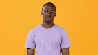 Genç bir adam mor gömlekli tarafsız bir yüz ifadesiyle kameraya düşünceli bir şekilde bakıyor. Canlı turuncu arka plan tavırlarına zıtlık katıyor..