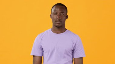 Mor bir gömlek giymiş, kendine güvenen genç bir adam, parlak turuncu bir arka planla çarpıcı bir kontrast yaratıyor. İfadesi ciddi, sakin tavırlarına dikkat çekiyor..