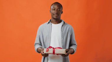 Genç bir adam elinde kırmızı kurdeleyle süslenmiş güzel paketlenmiş bir hediye kutusunu tutarken gülümsüyor parlak turuncu bir arka plana karşı neşeli bir beklenti sergiliyor..