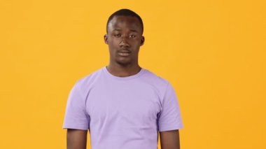 Genç bir adam, parlak sarı bir arka planın önünde, düz mor bir gömlek giyerek, tarafsız bir yüz ifadesi sergileyerek, sakin bir tavır sergiliyor..