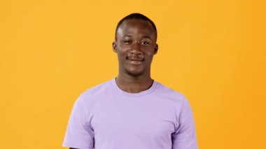 Neşeli genç bir adam açık mor bir tişört giymiş, geniş bir gülümsemeyle kendine güvenerek ayakta duruyor. Parlak turuncu arka plan neşeli atmosfere canlı bir dokunuş ekler..