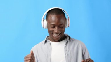 Genç bir adam kulaklık takıp gülümseyerek neşeyle dans ediyor. Parlak mavi arka plan, müzik zevkinin canlı atmosferini arttırır..