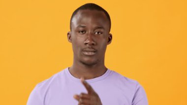 Genç bir adam, sessizliği simgelemek için işaret parmağını dudaklarına dayayarak güvenle ayakta duruyor. Açık mor bir gömlek giyer ve parlak sarı bir arka plana sahiptir..