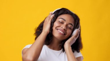 Genç, kıvırcık saçlı, kulaklık takmış, müzik zevkine dalmış, geniş bir şekilde gülümseyen siyah bir kadın. Canlı sarı arkaplan onun neşeli ifadesini ve pozitif ruh halini arttırır..
