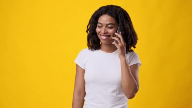 Kıvırcık saçlı genç bir Afrikalı Amerikalı kadın telefonda konuşurken parlak bir şekilde gülümsüyor. Neşeli sarı arka plan, sohbete neşe katıyor..