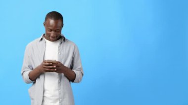 Açık tişört ve tişört giymiş genç bir adam akıllı telefonuna odaklanmış durumda. Dijital etkileşime dalmış, parlak mavi bir arkaplanın önünde duruyor..