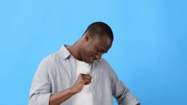 Bir adam aşağıya bakıyor, canlı mavi bir arkaplanın önünde dururken gömleğini düzeltiyor. Giysilerine odaklanmış görünüyor, sıradan bir tavırla..