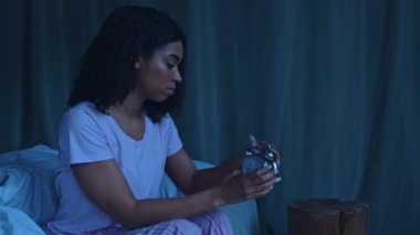 Pijamalı Afro-Amerikalı bir kadın sabah ışığında yatağında otururken çalar saati inceliyor..