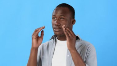 Acılı bir ifadeye sahip bir adam, yüzü parlak mavi bir arka plana dönük bir şekilde, hayal kırıklığı içinde başını sıkıca tutar. Yüz ifadesi stres ya da sıkıntı hissi veriyor..
