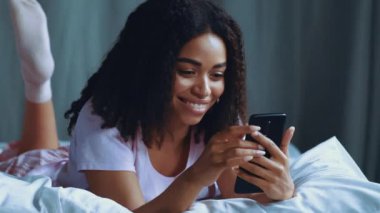Genç siyah bir kadın, rahat bir yatak odasında akıllı telefonunu kontrol ederken gülümseyerek yüzüstü yatıyor..