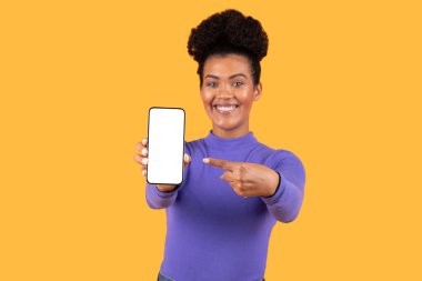 Kıvırcık saçlı, mor bluzlu bir kadın heyecanlı bir şekilde boş bir akıllı telefon ekranını işaret ediyor, canlı, sarı bir arka plana kurulmuş, coşkulu bir ifade veriyor..