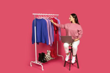 Taburede oturan bir kadın, dizüstü bilgisayarda çalışırken askıda giysileri inceliyor. Canlı arkaplan görüntülenen giysinin parlak renklerini güçlendirir ve canlı bir atmosfer oluşturur..