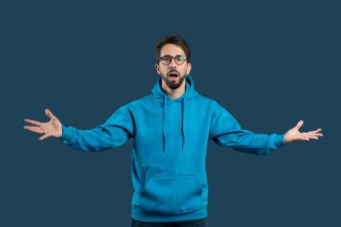 Mavi kapüşonlu genç bir adam kolları açık duruyor, koyu mavi bir arkaplana karşı sürpriz ya da coşku taşıyor. Yüzündeki ifade gerçek bir heyecanı yakalıyor..