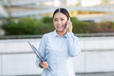 Asyalı kadın dışarıda duruyor, telefonda konuşurken gülümsüyor ve elinde bir yığın belge var. Arka planda, profesyonel bir ortam izlenimi veren yeşillik ve modern mimari yer alıyor..