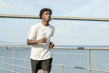 Açık tişörtlü ve şortlu genç bir Afro-Amerikalı, arka planda büyük bir köprü ile rıhtım yolu boyunca koşu yapar. Parlak gün ışığı dingin atmosferi güçlendirir..