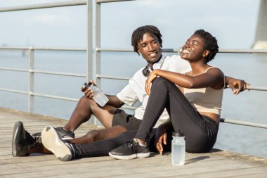 Afro-Amerikalı neşeli çift ahşap bir iskelede oturup, gülücüklerini ve içkilerini paylaşıyorlar. Sakin suyun yanında güneşli bir günün tadını çıkarıyorlar. Spor kıyafetlerinde sıcaklık ve bağlantı yayıyorlar..