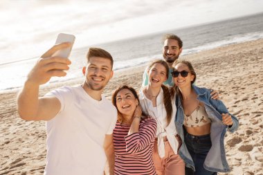 Bir grup arkadaş sahilde selfie çekerken mutlu bir şekilde poz veriyor. Mükemmel bir günde arka planda güneşin ve okyanus dalgalarının tadını çıkarıyorlar..