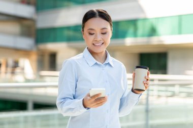 Genç bir Asyalı kadın dışarıda modern bir şehir bölgesinde duruyor, telefonuna mesaj atarken gülümsüyor ve elinde bir kahve fincanı tutuyor, güneşli bir anın tadını çıkarıyor..