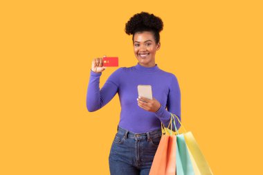 Genç bir kadın bir elinde kredi kartı, diğer elinde akıllı bir telefon, diğer kolundan sarkan parlak renkli alışveriş torbaları, alışveriş deneyiminde neşe sergiliyor..