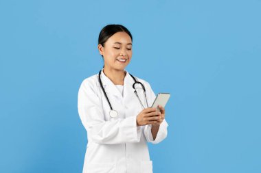 Beyaz önlük giymiş profesyonel bir Asyalı kadın akıllı telefonuyla mutlu bir şekilde meşgul oluyor. Parlak mavi bir ortamda modern teknoloji ve tıbbi uygulamaların bir karışımını sergiliyor..