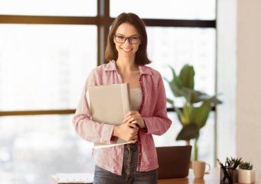 Kendine güvenen bir yönetici. Elinde dosyayla gülümseyen kadın ofis çalışanı. Kameraya bakıyor.