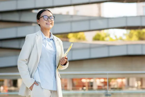 stock image A confident Asian woman dressed in a light suit stands outdoors, smiling as she holds a document. The modern architecture in the background enhances her professional demeanor, copy space