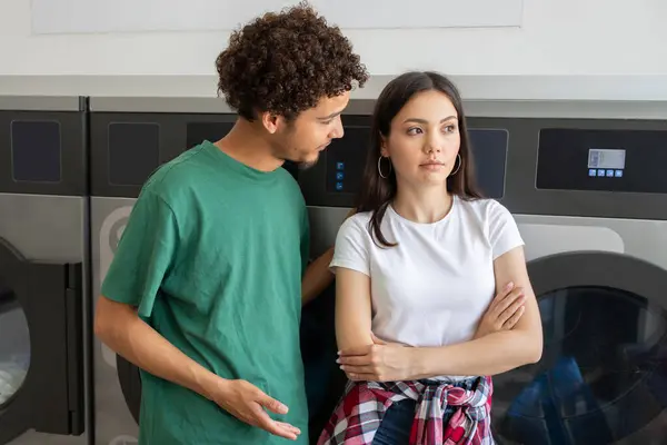 Stock image A young man and woman stand in a laundromat. The man appears to be discussing something serious with the woman, who looks thoughtful and slightly distressed while crossing her arms.