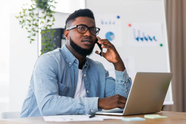 Genç Afrikalı Amerikalı yönetici dizüstü bilgisayarla çalışıyor ve endişeli bir yüz ifadesiyle telefonda konuşuyor, işte zor bir gün geçiriyor.