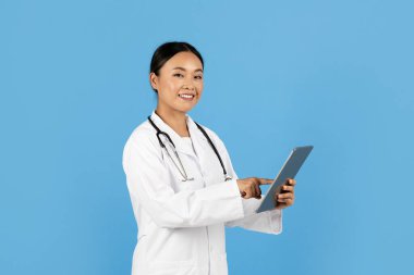 Gülümseyen Asyalı bir kadın sağlık uzmanı hasta bilgilerini kontrol etmek için tablet kullanarak mavi bir arkaplan karşısında duruyor. Boynunda steteskop olan beyaz bir ceket giyiyor..